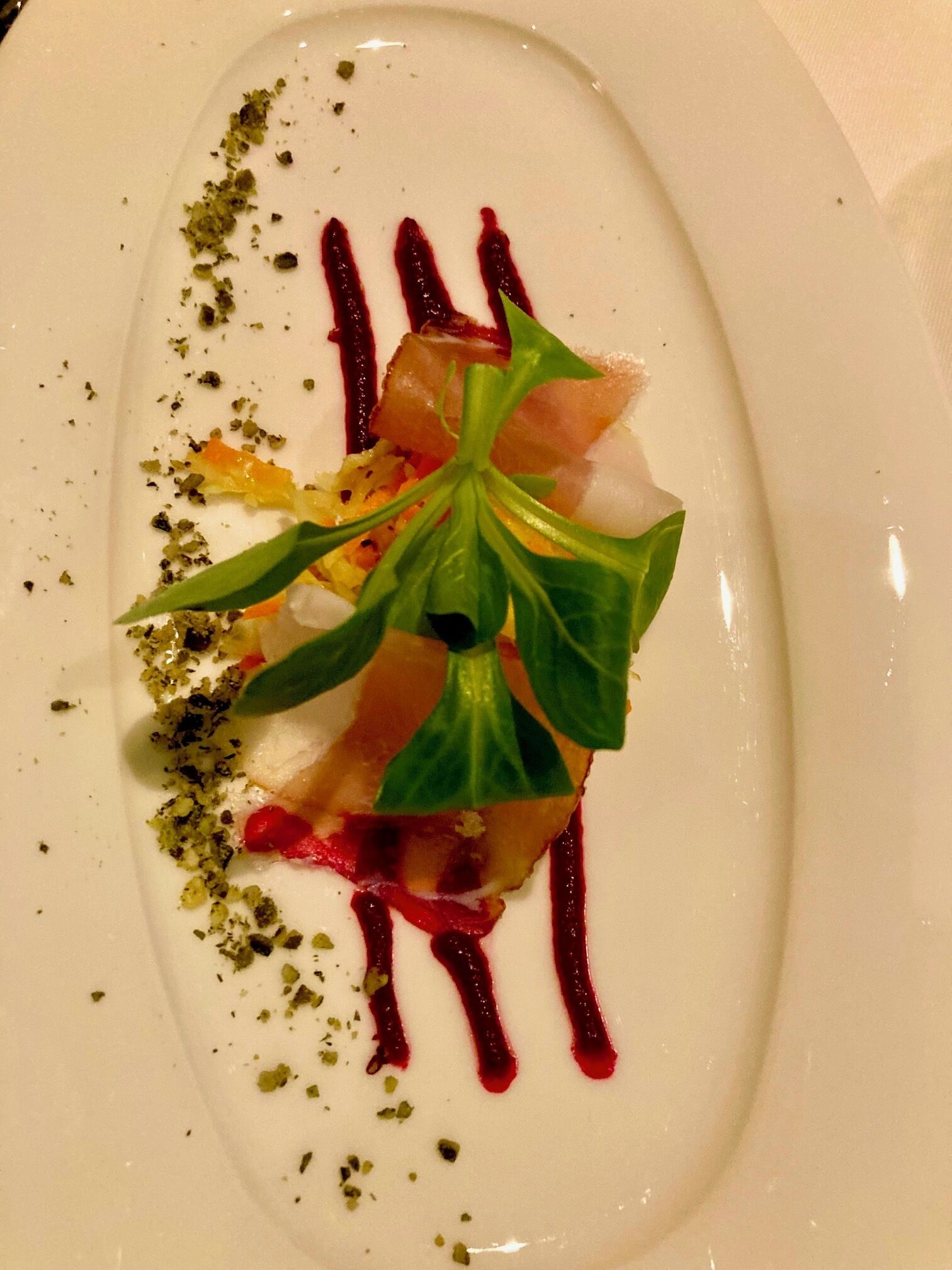 Une assiette blanche contenant un plat composé d’une grande feuille verte au centre, probablement du basilic, entourée d’une substance blanche et crémeuse, qui pourrait être une soupe ou une purée, le tout nappé d’une sauce rouge et parsemé d’herbes vertes.