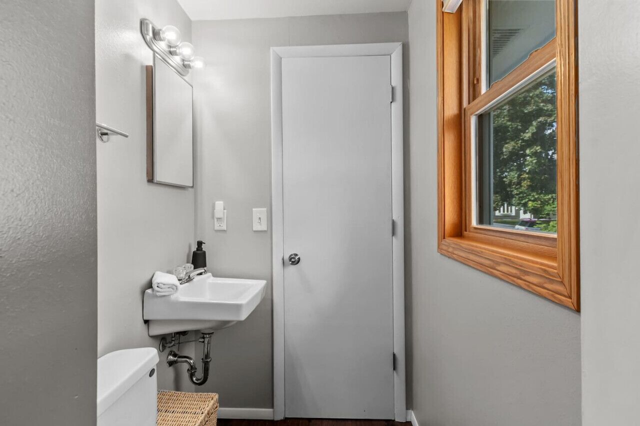 Een kleine badkamer met een witte wastafel, een spiegel met verlichting, een deur en een raam met uitzicht op groen.