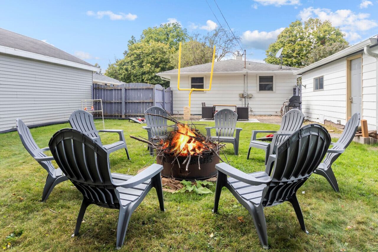 Un jardin avec un foyer entouré de quatre fauteuils Adirondack, un petit abri de jardin et une maison avec un garage.