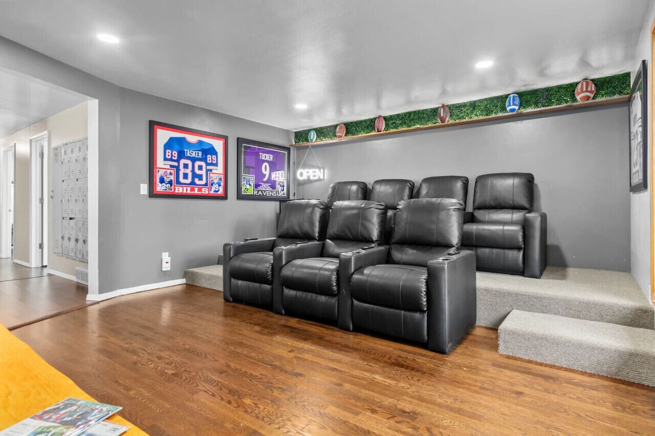 Une salle de cinéma maison moderne, dotée d’un grand ensemble de fauteuils inclinables en cuir noir, de murs gris et d’une décoration sur le thème du sport, comprenant des maillots de sport encadrés et une étagère avec des ballons de football.