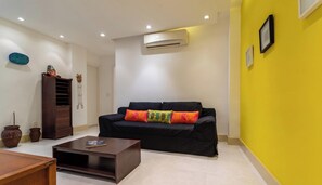 A living room with a black sofa, colorful cushions, a wooden coffee table, a wall-mounted air conditioner, and a yellow accent wall.