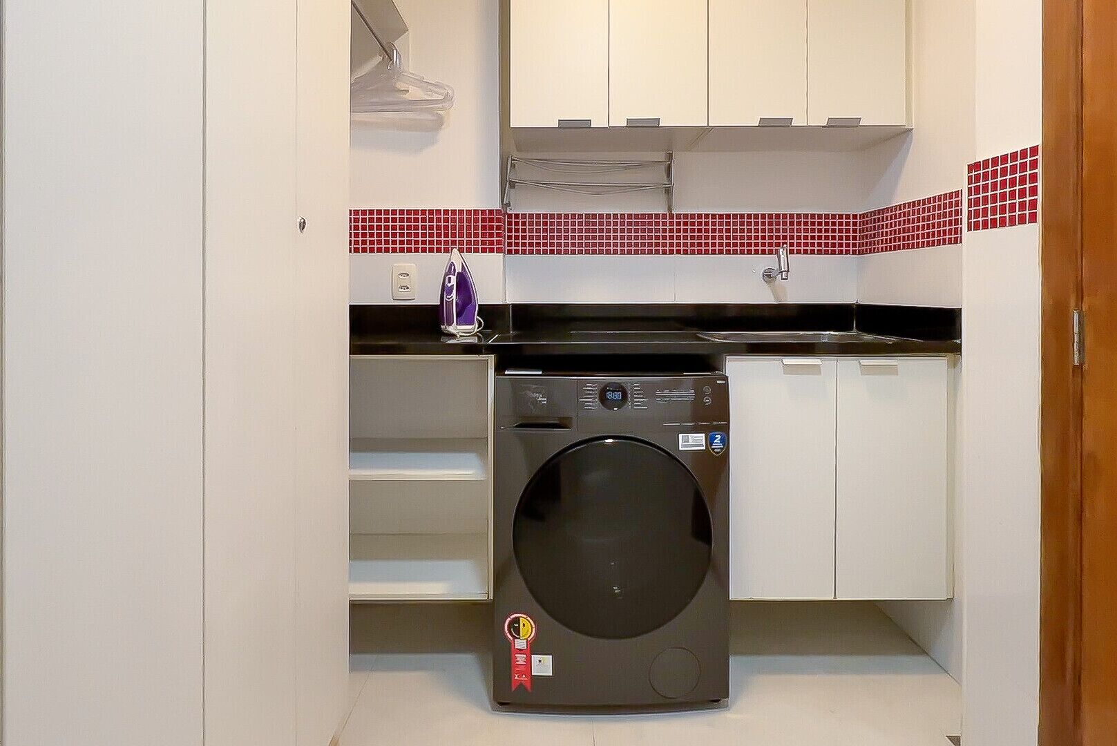 Ein kleiner Waschraum mit einer Frontlader-Waschmaschine, weißen Schränken und einer roten Fliesenrückwand.