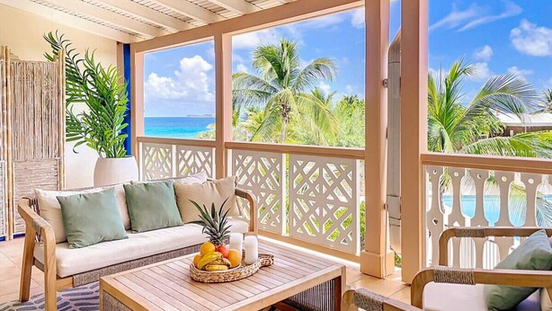A sunlit room with a wooden ceiling, a sofa with cushions, a coffee table with fruit, and a view of palm trees and the ocean.