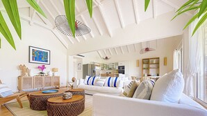 A modern living room with a white sofa, two round wooden ottomans, a coffee table, and a dining area visible in the background.