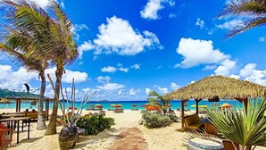 A beach pathway leading to a beach area with umbrellas, palm trees, and a thatched structure.