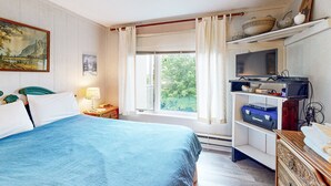 A bedroom with a bed, a television, a shelf with various items, and a window with curtains. - Cozy Mountain Retreat: Single-Level Condo in Sugar Mountain with Modern Comforts (Sugar Mountain)