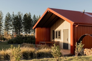 Une cabane en bois avec un toit rouge, une porte et une fenêtre. Il y a un banc à l’extérieur et une pile de bois de chauffage sur la droite.