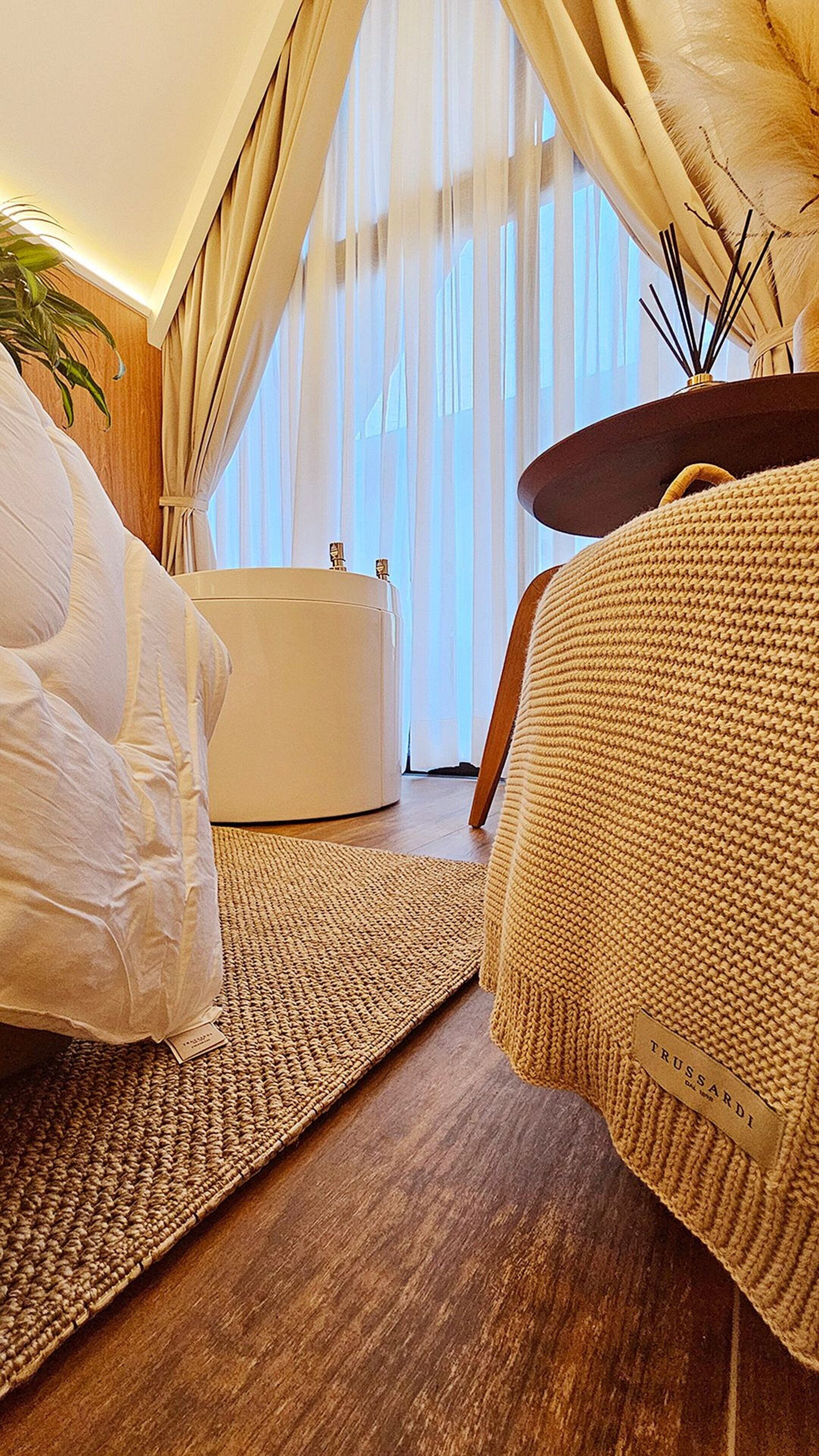 Ein gemütliches Hotelzimmer mit Holzboden, einem Webteppich, einem weißen Ottomanen, einem Webstuhl-Stuhl und Blick ins Freie durch transparente Vorhänge.
