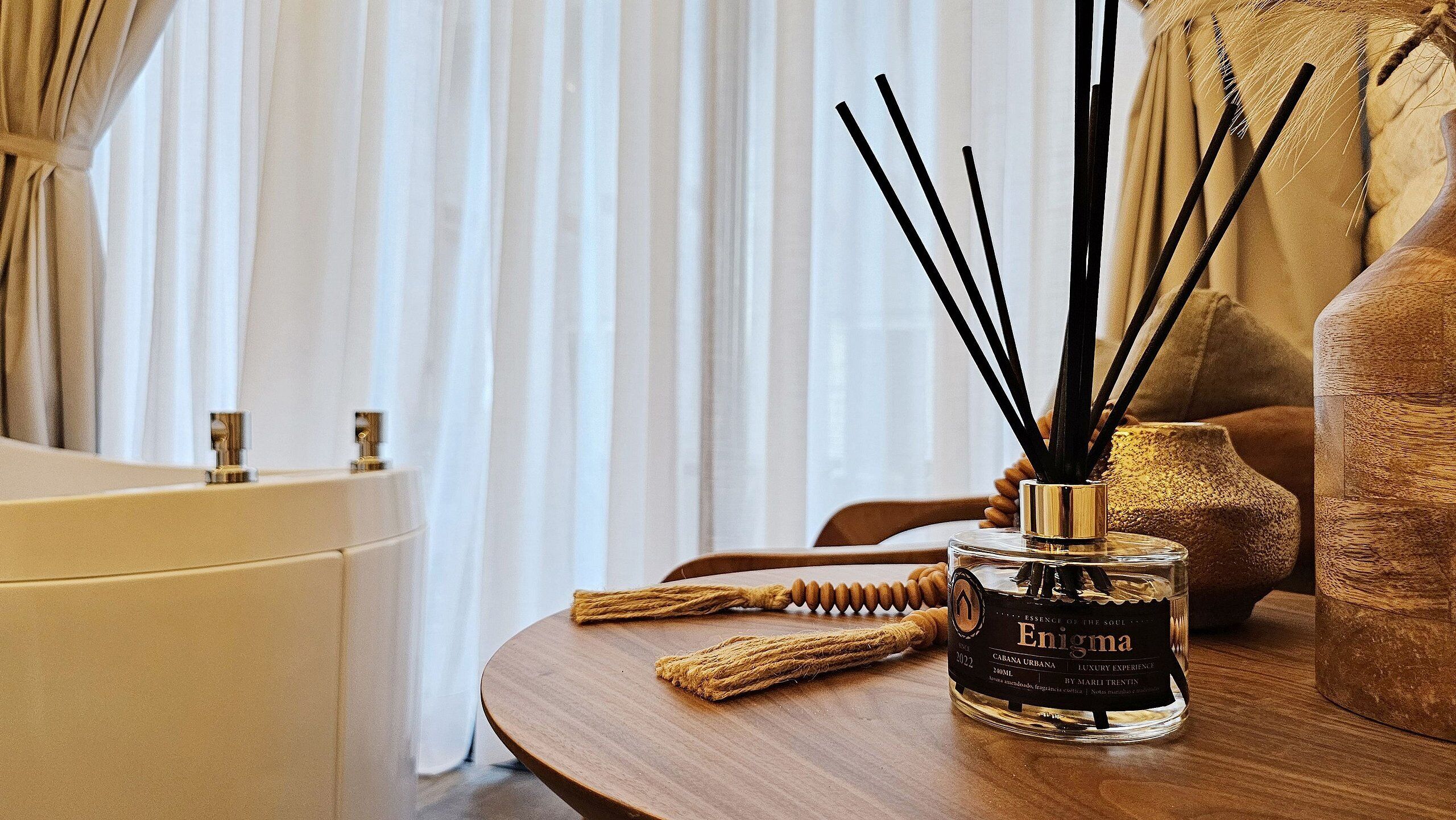 A bottle of Enigma cologne on a wooden table, a bathtub, and a window with curtains.