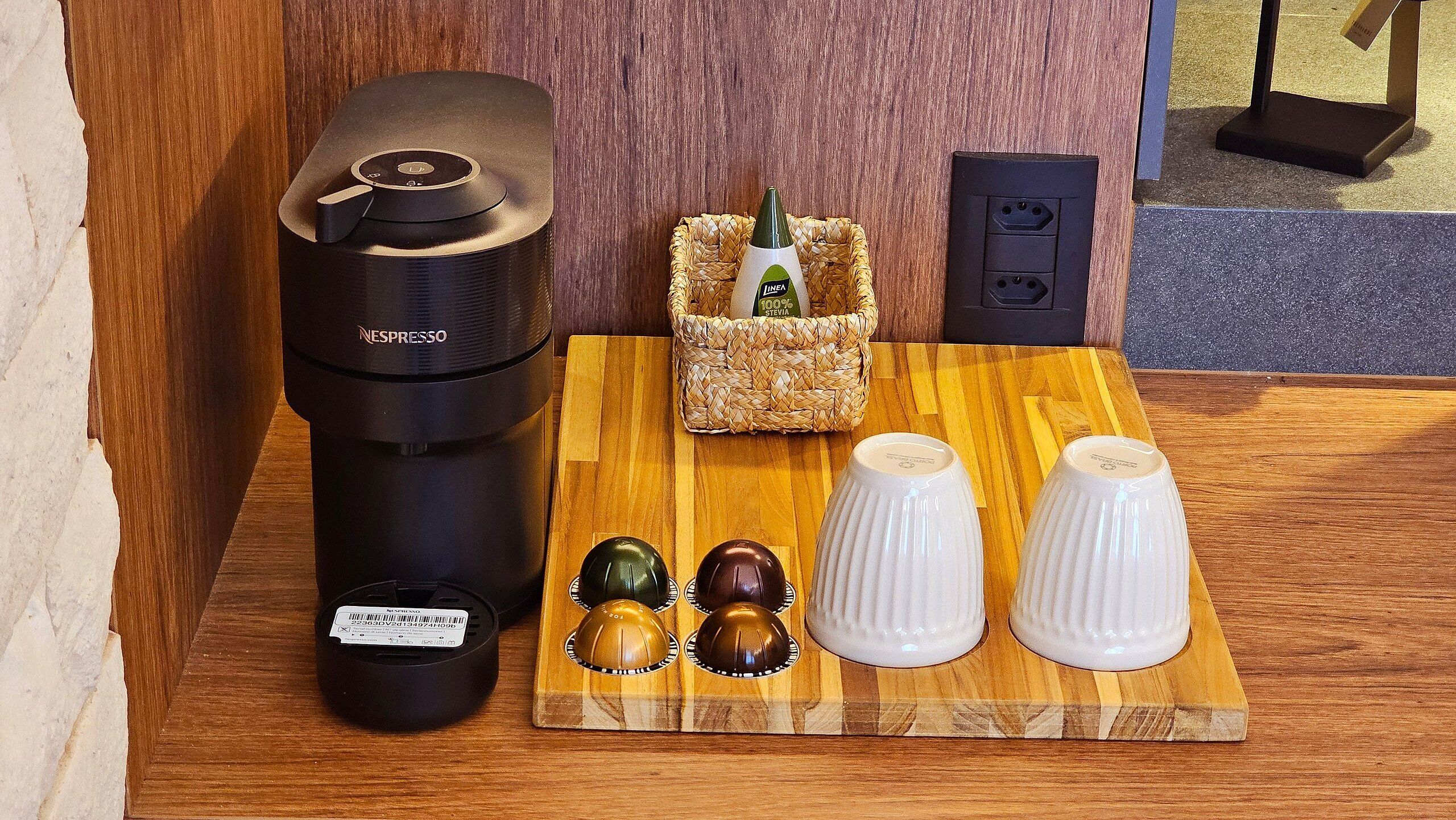 Una máquina de café Nespresso, una cesta con una botella y dos conjuntos de recipientes para condimentos sobre una mesa de madera.