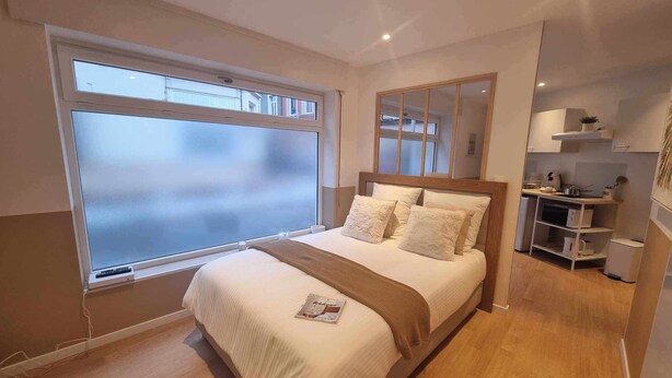 A modern bedroom with a large window, a bed with white bedding, a wooden headboard, and a small kitchenette area. - The White cosy, maxi studio in the city center, 1 km from the train station, tourism/business (Calais)