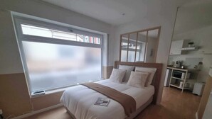 A modern bedroom with a large window, a bed with white bedding, a wooden headboard, and a small kitchenette area. - The White cosy, maxi studio in the city center, 1 km from the train station, tourism/business (Calais)
