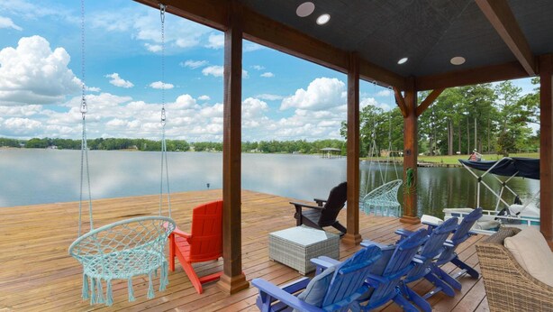 A covered deck with wooden flooring, outdoor seating including chairs and a hammock, and a view of a lake with boats and trees in the background.