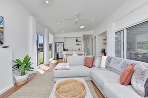 A modern living room with a sectional sofa, a wooden coffee table, and a view of the kitchen and outdoor area. - Magpie 2 3 Two for Joy Estate. 3 bedroom (North Rothbury)