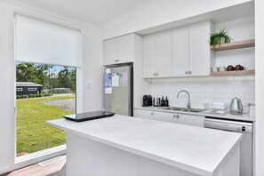 A modern kitchen with white cabinetry, a built-in dishwasher, and a window overlooking a grassy area. - Magpie 2 3 Two for Joy Estate. 3 bedroom (North Rothbury)