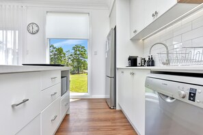 A modern kitchen with white cabinetry, a stainless steel refrigerator, a Bosch dishwasher, and a view of a green landscape through an open door. - Magpie 2 3 Two for Joy Estate. 3 bedroom (North Rothbury)