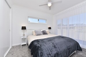 Une chambre à coucher comprenant un lit, deux tables de chevet, un ventilateur de plafond, une fenêtre avec des stores et une vue sur l’extérieur.