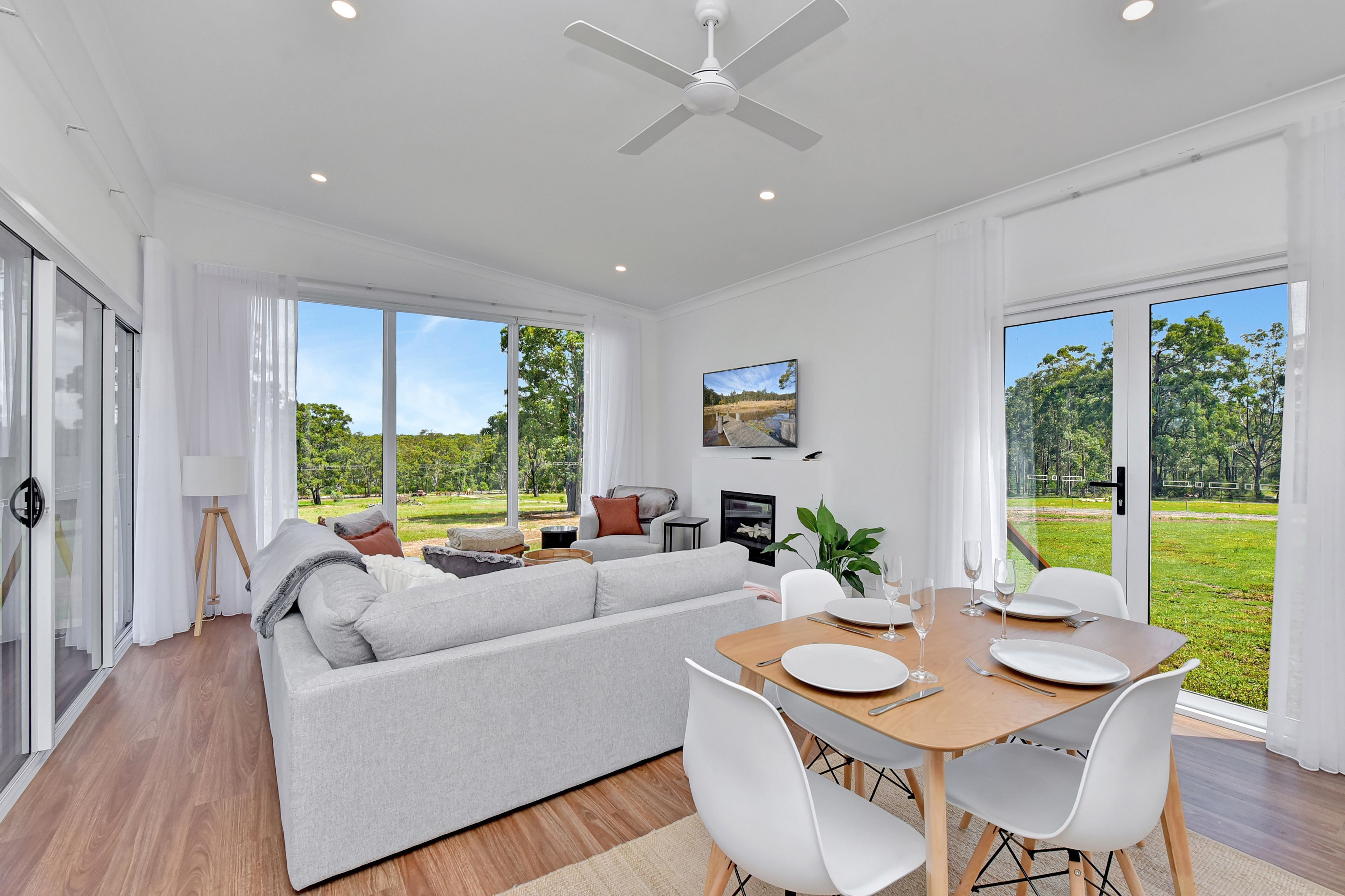 Un salon moderne comprenant un canapé, une table à manger et des chaises, de grandes fenêtres donnant sur un paysage verdoyant et un ventilateur de plafond.