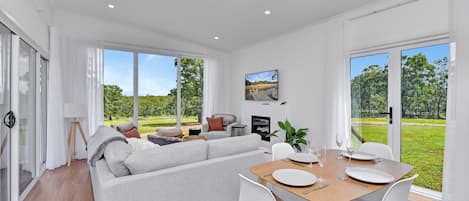Un salon moderne comprenant un canapé, une table à manger et des chaises, de grandes fenêtres donnant sur un paysage verdoyant et un ventilateur de plafond.