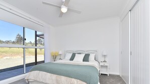 A bedroom with a large window, a bed with a white headboard, a nightstand, and a ceiling fan. - Magpie 2 3 Two for Joy Estate. 3 bedroom (North Rothbury)