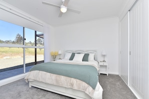 A bedroom with a large window, a bed with a white headboard, a nightstand, and a ceiling fan.