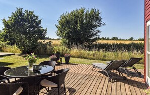 Une terrasse en bois, aménagée avec du mobilier d’extérieur, notamment une table à plateau en verre et des chaises en osier, donnant sur un champ et des arbres.