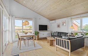 Een ruime woonkamer met een houten plafond, een eettafel met stoelen, een bank en een aan de muur bevestigde televisie.