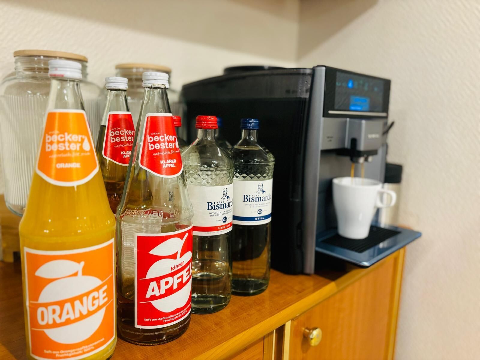 A variety of beverages including orange juice, apple juice, and water bottles on a wooden counter with a coffee machine in the background.