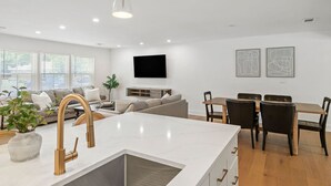 Een moderne keuken met een wit aanrecht en een gouden kraan, een eethoek met een houten tafel en stoelen, en een woonkamer met een bank en een televisie aan de muur.