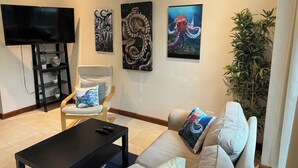 Un salon doté d’un téléviseur à écran plat, d’une table basse noire, d’un canapé beige agrémenté de coussins colorés, d’une chaise en bois et de trois œuvres d’art encadrées accrochées au mur.