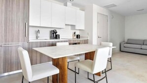 Una cocina moderna con armarios blancos, encimera de mármol y mesa de madera. Hay cuatro sillas blancas alrededor de la mesa.