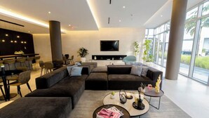 A modern living room with dark sofas, a coffee table, and a TV. There is a bar area with chairs and a large window providing natural light. - Modern Design District Condo, Balcony, Pool, Gym (Miami)
