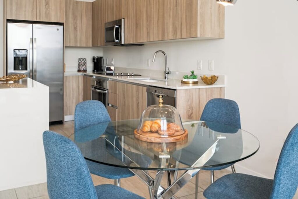 Una cocina moderna con una isla central, electrodomésticos de acero inoxidable y sillas azules alrededor de una mesa de vidrio.