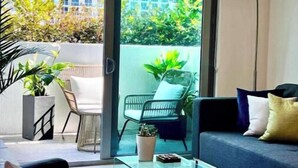 A modern living room with a glass coffee table, a sofa, and a chair. There is a view of a balcony with plants and a building in the background. - Relax with Pool, Gym, Balcony in Design District (Miami)