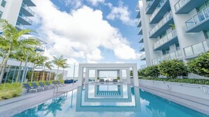 Ein modernes Hotel mit zentralem Pool, umgeben von Palmen und blauem Himmel.