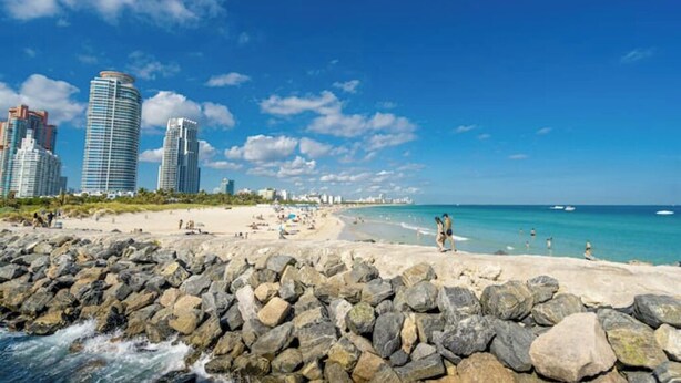 Ein Strand mit felsiger Küste, blauem Himmel und hohen Gebäuden im Hintergrund.