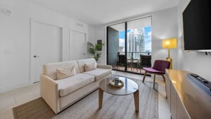 Ein modernes Wohnzimmer mit Sofa, Couchtisch und Fernseher. Ein Balkon bietet Blick auf Gebäude.