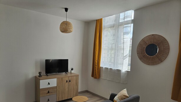 En moderne stue med et fladskærms-tv på et træskab, en grå sofa, et lille rundt bord og et stort vindue med lette gardiner.