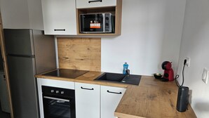 Et kompakt køkken med hvide skabe, et rustfrit stål køleskab, indbygget mikrobølgeovn, opvaskemaskine og en træbordplade.