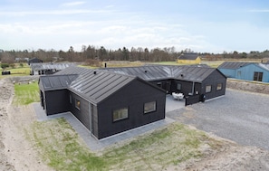 Un bâtiment moderne à l’extérieur sombre et doté d’un toit en pente, entouré d’une zone recouverte de gravier et d’un champ d’herbe.