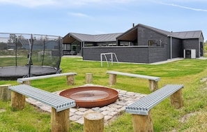 Une maison moderne dotée d’un grand jardin, comprenant un trampoline, un foyer extérieur et un espace patio avec des bancs.