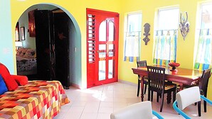 A room with a bed, a dining table with chairs, a red door, and a window with curtains.