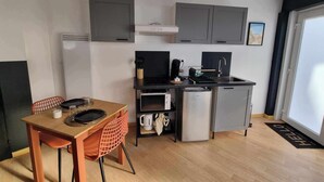 A compact kitchen with grey cabinets, a small refrigerator, and a coffee machine. There is a dining table with a chair and a mat on the floor. - The black tendance, maxi studio hyper center, 1km from train station, tourism/business (Calais)