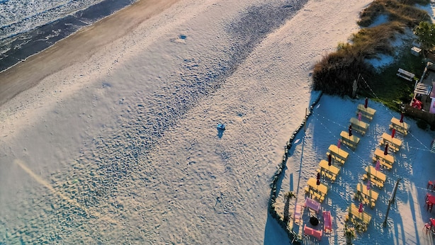 An aerial view of a beach with a pier extending into the water, clear skies, and a calm sea.