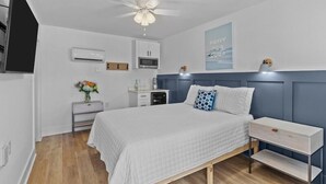 A bedroom with a bed, bedside tables, a ceiling fan, an air conditioning unit, a microwave, a small cabinet, and a decorative wall piece.