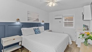 Un dormitorio con cama, mesitas de noche, ventilador de techo, equipo de aire acondicionado, ventana con persianas y un escritorio pequeño con un florero.