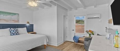 Ein Schlafzimmer mit einem Bett, einem Nachttisch, einem Deckenventilator, einer Tür zu einem Balkon und einer kleinen Küchenzeile mit Arbeitsfläche und einer Flasche.