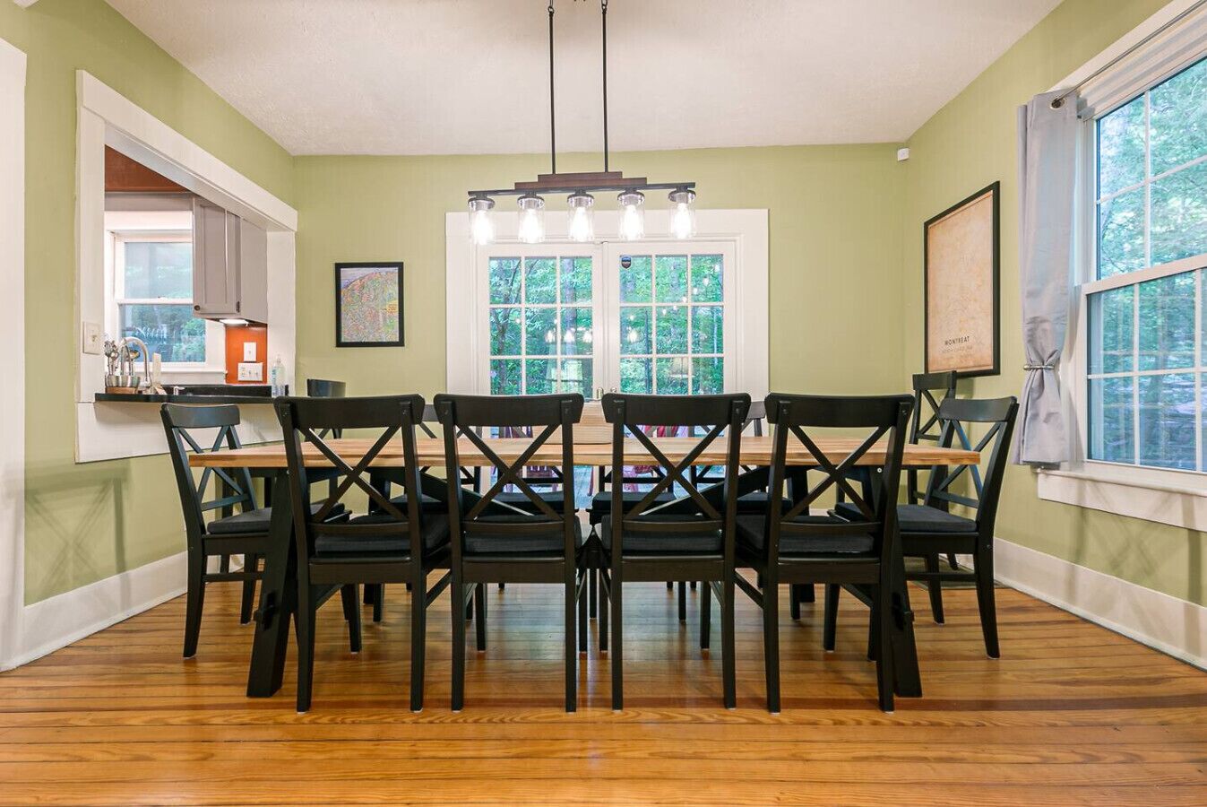 A dining room with a wooden table and black chairs, large windows, and a kitchen area visible through an open door.