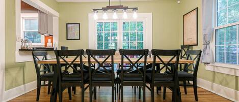 A dining room with a wooden table and black chairs, large windows, and a kitchen area visible through an open door.