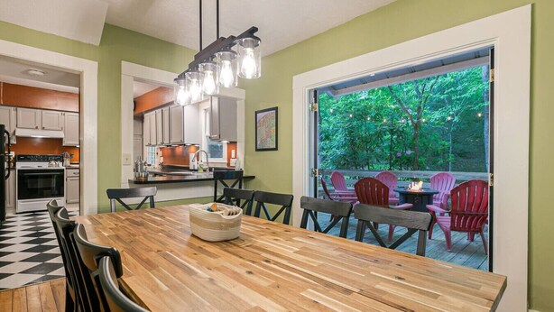 A dining area with a wooden table and chairs, a kitchen in the background, and a view of a deck with outdoor seating.
